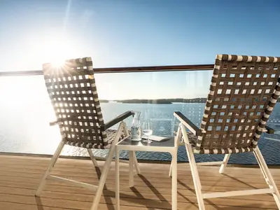 Zwei moderne Klappstühle und ein kleiner Tisch mit Wasserkaraffe und Gläsern auf einem Balkon der Mein Schiff 2. Im Hintergrund ist das glitzernde Meer mit kleinen Inseln bei Sonnenuntergang zu sehen.