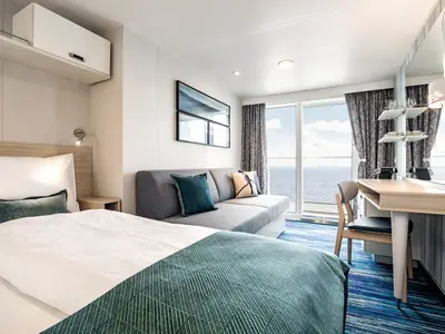 Ein helles Schlafzimmer auf der Mein Schiff 2 mit einem Bett im Vordergrund und einem Sofa in der Mitte. Im Hintergrund ist ein Balkon mit Meerblick zu sehen. Ein Schreibtisch mit Spiegel befindet sich rechts.
