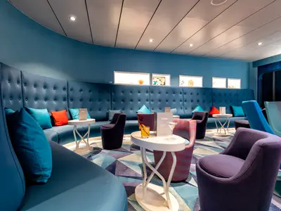 Innenansicht der TUI Bar an Bord der Mein Schiff 2, geprägt von tiefblauen Polstermöbeln, gemustertem Teppich und kleinen Tischen mit Getränken. Die Wand ist mit beleuchteten Nischen und Kunstobjekten verziert. Verschiedene Sessel in Lila, Rot und Türkis laden zum Verweilen ein.