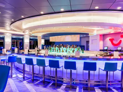 Innenansicht der TUI Bar auf der Mein Schiff 2: Eine modern beleuchtete, halbrunde Bar mit einem hellblauen Tresen und blauen Barhockern davor. Hinter dem Tresen stehen auf mehreren beleuchteten Ebenen zahlreiche Spirituosenflaschen. Im Hintergrund ist an der Wand ein großes, rotes TUI-Lächeln-Logo zu sehen, daneben zwei Bildschirme an einem Servicetresen.