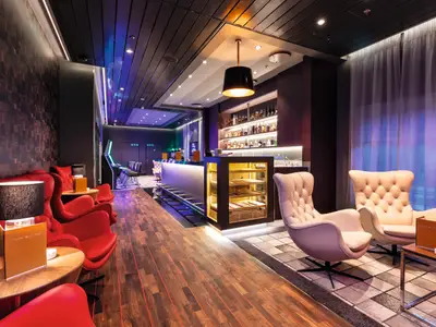 Eine moderne Bar und Lounge an Bord der Mein Schiff 2. Links stehen rote Sessel an Holztischen, rechts helle Ledersessel auf einem gemusterten Teppich. Im Hintergrund befindet sich eine beleuchtete Bar mit Alkoholregalen und Spielautomaten. Die Decke ist dunkel mit Einbauleuchten.