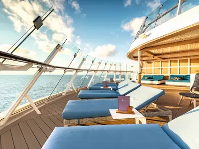 Ein helles Pooldeck an Bord der Mein Schiff 2 mit mehreren Reihen blauer Liegestühle aus Rattan und kleinen weißen Beistelltischen. Im Hintergrund ist das blaue Meer zu sehen, darüber ein sonniger Himmel mit weißen Wolken. Eine gebogene Glasbalustrade trennt das Deck vom Meer. Im rechten Bildbereich sind Sitzecken mit türkisfarbenen Polstern und eine dunkle Korbstruktur zu erkennen.