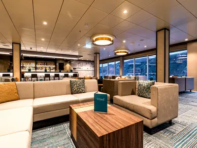 Ein breiter Blick in die Ruhepol Bar & Lounge an Bord der Mein Schiff 2. Bequeme beigefarbene Sofas und Sessel stehen auf einem Teppich mit geometrischem Muster. Im Hintergrund eine lange Bar und große Fenster mit Blick auf eine bergige Küste.