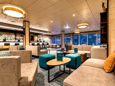 Ein geräumiger und modern eingerichteter Loungebereich an Bord der Mein Schiff 2. Links eine voll ausgestattete Bar mit vielen Flaschen. Im Vordergrund bequeme Sofas und Hocker in Grau und Türkis, mit kleinen Tischen. Große Fensterfronten bieten einen Blick auf eine blaue Landschaft. Deckenleuchten spenden warmes Licht.