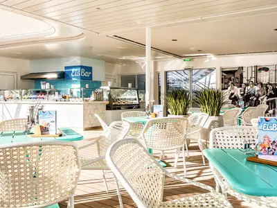 Innenansicht der Eisbar "EisBar by Bruno Gelato" an Bord der Mein Schiff 2. Helle, moderne Einrichtung mit vielen weißen Rattanstühlen und mintgrünen Tischen. Eine lange Theke mit Eisvitrinen ist zu sehen. Eine große Wandgrafik zeigt Menschen, die Eis essen. Große Fensterfronten bieten Meerblick.