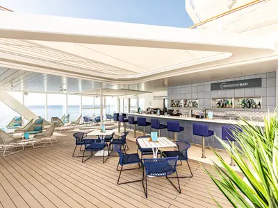 Blick in die helle Unverzichtbar-Lounge an Bord der Mein Schiff 2. Links sind gemuetliche Sessel mit Meerblick zu sehen, rechts eine Bar mit blauen Hockern.