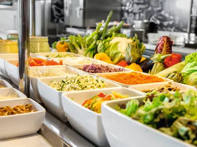 Eine gut gefüllte Salatbar auf der Mein Schiff 2, mit einer Vielzahl von frischen Zutaten in weißen Schalen. Man sieht verschiedene Salatsorten, geraspelte Karotten, Zucchini, Rotkohl, Pilze, Nüsse und Samen. Im Hintergrund sind frisches Gemüse und Früchte, wie Paprika, Artischocken, Auberginen und ganze Salatköpfe zu erkennen. Die Beleuchtung ist hell und einladend, und der Fokus liegt auf den Essensoptionen.