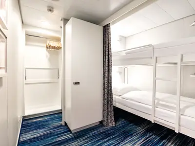 Blick in eine moderne, weiß eingerichtete Kabine an Bord der Mein Schiff 2. Links ein offener Kleiderschrank mit Holzkleiderbügeln, rechts Etagenbetten mit weißer Bettwäsche und einer Leiter. Ein gemusterter Vorhang trennt den Bereich der Betten ab. Der Boden ist mit einem blauen Teppich ausgelegt.