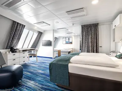 Innenansicht einer geräumigen, modernen Kabine der Mein Schiff 2. Links große Fenster mit Blick auf das Meer, davor ein Schreibtisch mit Kaffeemaschine und Schubladen. Rechts ein Doppelbett mit teal-farbener Tagesdecke. Im Hintergrund ein zusätzliches Einzelbett. Der Teppich ist blau-weiß gestreift. An der Wand hängt ein Fernseher.
