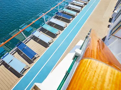 Blick von oben auf das Deck der Mein Schiff 2. Links das tiefblaue Meer, rechts eine Reihe blauer Liegestühle entlang einer blauen Laufbahn mit weißer Linie. Rechts ein hölzernes Geländer mit Reflexionen.