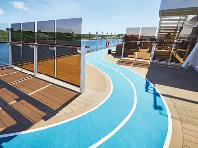 Sonniges Außendeck der Mein Schiff 2 mit einer geschwungenen, blauen Joggingstrecke. Rundum Glasgeländer mit Holzhandläufen. Im Hintergrund ist das Meer und eine grüne Küstenlandschaft zu sehen. Seitlich rechts führen Treppen nach oben.
