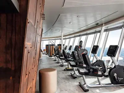 Ein langgezogener Fitnessraum auf der Mein Schiff 2 mit vielen Recumbent-Bikes und anderen Geräten, die entlang einer großen Fensterfront zum Meer ausgerichtet sind. Rechts im Bild sind einige der Recumbent-Bikes von Life Fitness zu sehen, die Bildschirme für Trainingsdaten haben. Die Wände auf der linken Seite sind in einem rostfarbenen, industriellen Stil gehalten. Der Boden ist hellgrau gefliest.