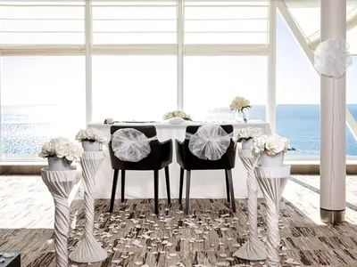 Festlich dekorierter Bereich an Bord der Mein Schiff 2 fuer eine Hochzeit. Zwei Stuehle, Tisch mit weissen Blumen und Rosenblaetter auf dem Boden. Heller Meerblick durch grosse Fenster.