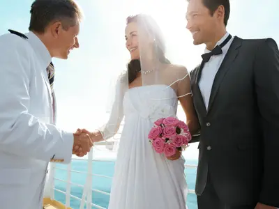 Ein lächelndes Brautpaar und ein Kapitän schütteln sich an Bord der Mein Schiff 2 auf dem Außendeck die Hände. Die Braut hält einen Strauß pinker Rosen.