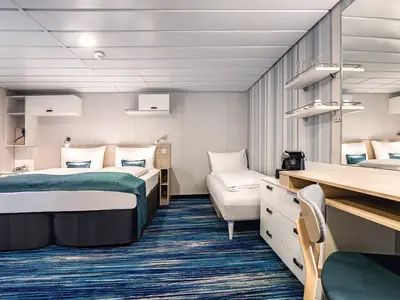 Innenansicht einer modern eingerichteten Kabine an Bord der Mein Schiff 2. Sie zeigt ein großes Doppelbett und ein Einzelbett, beide mit weißen Laken und blauen Kissen und Decken. Ein blau-gestreifter Teppich bedeckt den Boden. Rechts ist ein Schreibtisch mit Stuhl und ein Wandspiegel zu sehen. An der Wand hängen Regale und Bilder.
