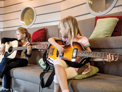 Zwei junge Mädchen spielen Gitarren auf einem Sofa im Kinderbereich "Insel der Seeräuber" an Bord der Mein Schiff 2. Das linke Mädchen spielt Akustikgitarre, das rechte Mädchen eine Bassgitarre. Sie schauen sich gegenseitig an.
