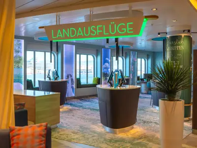 Blick in den Landausflugsbereich der Mein Schiff 2. Mehrere halbrunde Tresen mit Monitoren stehen vor grossen Fenstern, die den Blick auf eine Stadt am Hafen freigeben. Eine grosse Leuchtschrift in Gruen zeigt "LANDAUSFLÜGE".