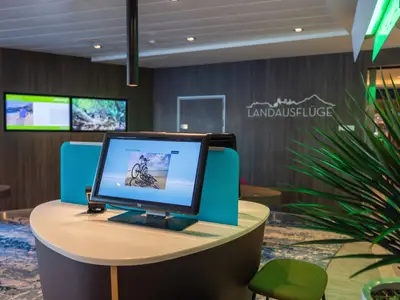 Ein Landausflug-Terminal mit Touchscreen und Fahrrädern am Strand als Motiv. Im Hintergrund weitere Bildschirme und die Aufschrift LANDAUSFLÜGE an Bord der Mein Schiff 2.