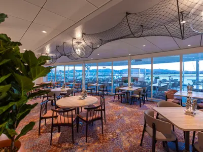 Innenansicht des Restaurants Fischmarkt an Bord der Mein Schiff 2 mit zahlreichen Tischen und Stühlen. Große Fensterfronten bieten Meerblick auf eine Stadt und Berge im Hintergrund. Decken sind mit Fischernetzen und Leuchten dekoriert.