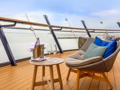 Gemütliche Sitzecke an Deck der Mein Schiff 2: Ein Korbsessel mit Kissen, Beistelltisch, Champagnerkühler, Flasche und Gläsern. Moderne Glasbrüstung und blauer Himmel im Hintergrund.