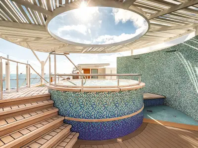 Blick auf den runden Whirlpool mit gemusterten Mosaikfliesen und die angrenzende Duschzone mit zwei Duschköpfen auf dem Sonnendeck der Mein Schiff 2. Der Boden ist mit hellem Holz verlegt, und eine Treppe führt zu einer Aussichtsplattform mit Glasgeländer. Im Hintergrund ist das Meer unter blauem Himmel mit Wolken zu sehen.