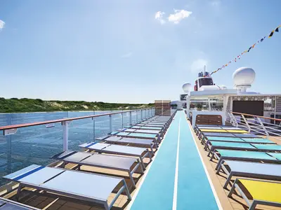 Blick auf das obere Außendeck der Mein Schiff 2. Links reihen sich leere Sonnenliegen und eine blaue Laufbahn. Rechts ist die Küste mit grünen Bäumen und Sand zu sehen. Im Hintergrund des Schiffes sind Schornsteine und Radarkuppeln erkennbar, davor bunte Flaggen. Der Himmel ist blau mit einigen Wolken.