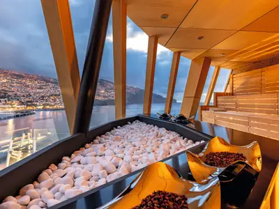 Ein luxuriöser Saunabereich an Bord der Mein Schiff 2 bei Sonnenuntergang. Durch große Fenster blickt man auf eine beleuchtete Küstenstadt und das Meer. Im Vordergrund liegen weiße Steine in einem Becken. Im Hintergrund befinden sich Saunabänke aus hellem Holz mit indirekter Beleuchtung. Eine goldene Schale mit getrockneten Blütenblättern steht auf einer schwarzen Ablage.