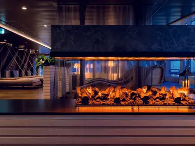 Ein moderner Elektrokamin mit leuchtenden Holzscheiten im Spa-Bereich der Mein Schiff 2, dahinter Spiegelungen von Entspannungsliegen in einer beruhigenden, dunkelblauen Umgebung.