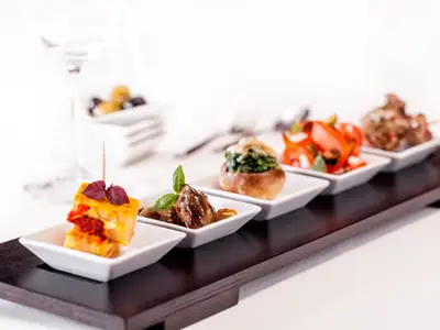 Fuenf verschiedene Tapas-Gerichte in kleinen weissen Schaelchen, arrangiert auf einem dunklen Holztraeger. Im unscharfen Hintergrund sind Weissweinglaeser und weitere Schalen der Mein Schiff 2 zu sehen.
