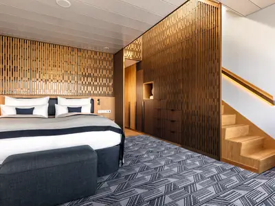 Blick in eine moderne Kabine der Mein Schiff 2 mit einem gemütlichen Doppelbett, einem geometrisch gemusterten Teppich und vertikalen Holzlamellen an den Wänden. Eine kleine Treppe führt nach oben.