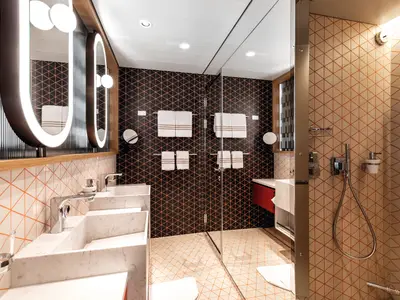Dieses Bild zeigt ein stilvolles Badezimmer der Mein Schiff 2. Es hat zwei ovale, beleuchtete Spiegel über Doppelwaschbecken. Die Wände sind mit dreieckigen Mustern in Schwarz und Orange, sowie Beige und Orange gefliest. Rechts befindet sich ein Duschbereich mit Glaswand und modernen Armaturen.