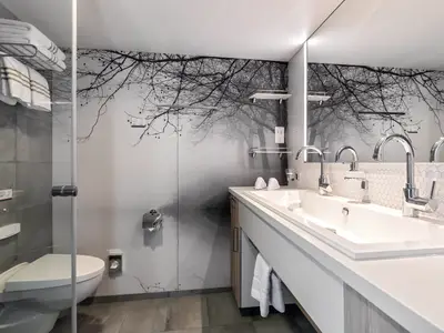 Ein modernes Badezimmer an Bord der Mein Schiff 2 mit einem Doppelwaschbecken, einer begehbaren Dusche und einer Schwarz-Weiß-Tapete, die einen nebligen Wald zeigt. Weiße Handtücher liegen bereit.