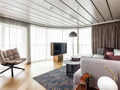 Eine helle, stilvolle Suite an Bord der Mein Schiff 2 mit raumhohen Fenstern, die einen weiten Blick bieten. Moderne Möbel, darunter ein bequemer Sessel, ein großer Flatscreen-Fernseher und ein Ecksofa mit verschiedenen Kissenfarben, sind auf dem Holzboden platziert. Der Raum ist mit Gardinen und einem gemusterten Teppich ausgestattet.