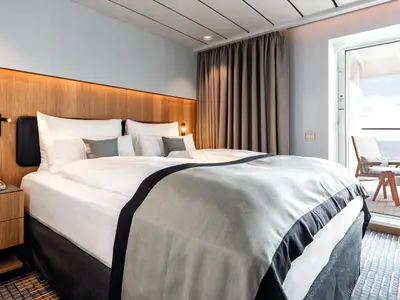 Ein komfortabel eingerichtetes Doppelzimmer an Bord der Mein Schiff 2 mit einem breiten Bett, weißen Laken und einer grauen Decke. An der Seite ist ein Holznachttisch mit Telefon. Im Hintergrund ist ein Balkon mit Meerblick zu sehen.
