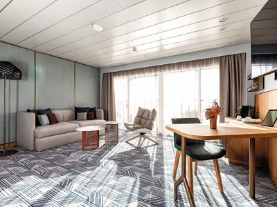 Ein heller, moderner Wohnbereich an Bord der Mein Schiff 2. Er zeigt ein Sofa mit Kissen, zwei Beistelltische, einen Sessel, einen Esstisch mit Stühlen und eine Kaffeemaschine. Tageslicht strömt durch große Fenster. Der Teppich hat ein blau-weißes geometrisches Muster.