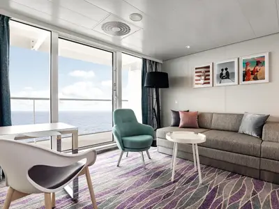 Ein helles Wohnzimmer mit großer Glasfront zum Meer auf der Mein Schiff 2. Links ein Schreibtisch mit Stuhl, mittig ein türkisfarbener Sessel, rechts ein graues Sofa mit Kissen und einem kleinen runden Beistelltisch. An der Wand hängen drei Bilderrahmen mit Bademodenmotiven.