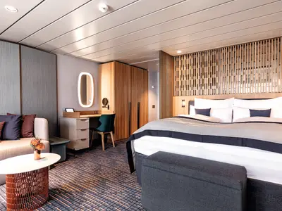 Blick in eine moderne Hotelkabine an Bord der Mein Schiff 2. Links ein helles Sofa mit Kissen, rechts ein großes Doppelbett mit weißer Bettwäsche und dunkelgrauer Decke. Im Hintergrund ein Schreibtisch mit Spiegel und ein Holzschrank.
