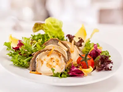 Nahaufnahme eines Salats auf einem weißen Teller, angerichtet mit grünem Blattsalat, runden Scheiben von gebackenem Ziegenkäse, dünnen Fleischscheiben, einem ganzen Cherrytomate mit Stiel und gelben sowie roten Salatblättern. Das Gericht wird in einem Restaurant der Mein Schiff 3 serviert.