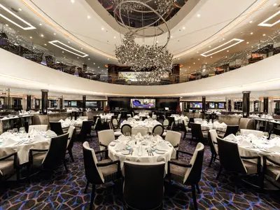 Blick in das weitläufige, elegante Restaurant der Mein Schiff 3. Zahlreiche runde Tische sind gedeckt, ein großer, kunstvoller Kronleuchter hängt in der Mitte. Eine obere Ebene mit weiteren Sitzplätzen ist sichtbar.