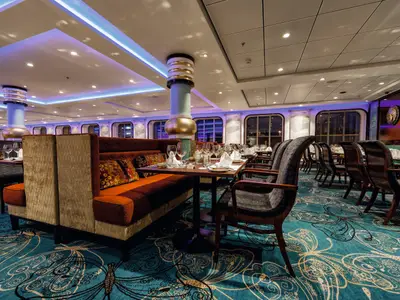 Innenansicht eines Restaurants auf der Mein Schiff 3. Elegante Sitzbänke und Tische sind für Gäste gedeckt. Blaue LED-Lichter zieren die Decke und ein gemusterter Teppich in Türkis schmückt den Boden. Große Fenster geben den Blick nach draußen frei.