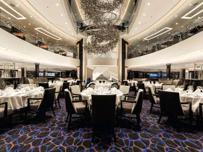 Blick in das elegant gedeckte Hauptrestaurant Atlantik Klassik an Bord der Mein Schiff 3. Zahlreiche Tische sind mit weißen Tischdecken und Geschirr ausgestattet. Im Hintergrund sind auf mehreren Ebenen weitere Bereiche des Restaurants und Bars zu sehen. Ein großer, kunstvoller Kronleuchter hängt von der Decke.