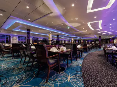 Ein elegantes Restaurant an Bord der Mein Schiff 3 bei Nacht. Violette und blaue Beleuchtung erhellt den Raum, der mit dunklen Holztischen und Stühlen mit Samtpolsterung ausgestattet ist. Der Teppich hat ein aufwendiges blaues und goldenes Muster. Gedeckte Tische mit weissen Servietten und Weingläsern sind bereit für Gäste. Ein runder Säulenakzent mit spiegelnden Oberflächen ist zu sehen.