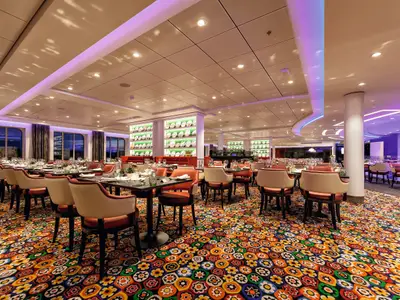 Innenansicht des Restaurants 'Atlantic Mediterran' auf der Mein Schiff 3. Elegante Tische sind für Gäste vorbereitet, umgeben von Stühlen mit cremefarbenen Lehnen und korallenroten Sitzflächen. Die Wände sind mit dekorativen Tellern in Regalen geschmückt, während der Boden ein auffälliges, farbenfrohes Blumenmuster aufweist. Große Fenster bieten einen Blick nach draußen. Die Decke ist hell und mit indirekter Beleuchtung sowie Spotlights versehen.