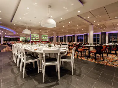 Blick in das elegant eingerichtete Hauptrestaurant Atlantik Klassik an Bord der Mein Schiff 3. Eine lange weiße Tafel mit weißen Stühlen dominiert den Vordergrund. Im Hintergrund sind weitere Tische mit roten Stühlen und große Fenster zu sehen. Eine Wanddekoration mit Tellern und eine lila Beleuchtung runden das Ambiente ab.