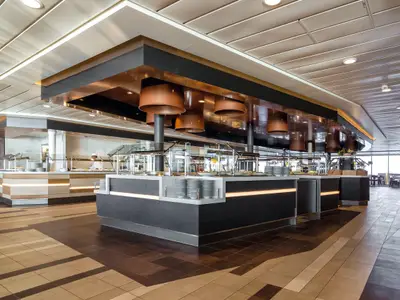 Innenansicht des Buffetrestaurants auf der Mein Schiff 3 mit einem langen, modernen Buffetbereich in dunklen und hellen Holztönen. Kupferfarbene Lampen hängen von der Decke. Ein Koch steht hinter einem Teil des Buffets. Im Hintergrund sind große Fenster und Tische zu sehen.