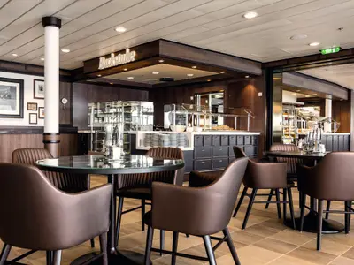 Ein warmer, einladender Speiseraum an Bord der Mein Schiff 3. Der Raum ist mit dunklem Holz vertäfelt und hat eine moderne Buffetstation mit frischen Speisen und glänzenden Glasvitrinen. Mehrere runde Tische mit bequemen braunen Stühlen stehen bereit. Ein Schild mit der Aufschrift "Backstube" leuchtet über dem Buffet.