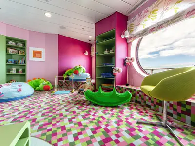 Ein farbenfrohes Spielzimmer an Bord der Mein Schiff 3 mit pinken und gruenen Waenden und einem grossen, runden Bullaugenfenster mit Blick auf das Meer und den Himmel. Ein gruener Sessel steht am Fenster, und auf dem gemusterten Teppichboden liegen Spielzeuge wie Stofftiere und eine gruene Wippe.