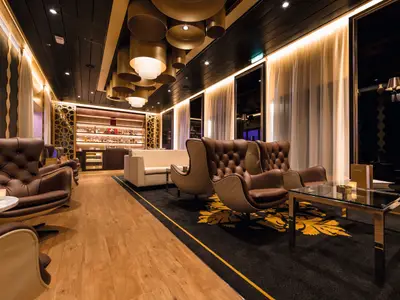 Blick in die elegante Bar der Mein Schiff 3. Braune Ledersessel und cremefarbene Loungesofas stehen auf hellem Holzboden und einem dunklen Teppich mit goldenem Muster. Im Hintergrund ist eine gut gefüllte Bar mit beleuchteten Regalen zu sehen, rechts daneben goldene Trennwände.