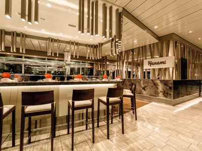 Innenansicht des japanischen Restaurants Hanami auf der Mein Schiff 3. Man sieht eine Bar mit mehreren Hochstühlen und ein beleuchtetes Sushi-Laufband. Im Hintergrund ist ein Schild mit der Aufschrift 'Hanami Japanische Küche' zu erkennen.