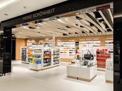 Detailansicht eines gut beleuchteten Duty-Free-Shops an Bord der Mein Schiff 3 mit vielen Regalen voller Kosmetikprodukte und Parfüms bekannter Marken wie Chanel, Dior und Gucci. Im Vordergrund sind Auslagen mit Handtaschen und weiteren Produkten zu sehen. Die Decke ist modern gewellt.